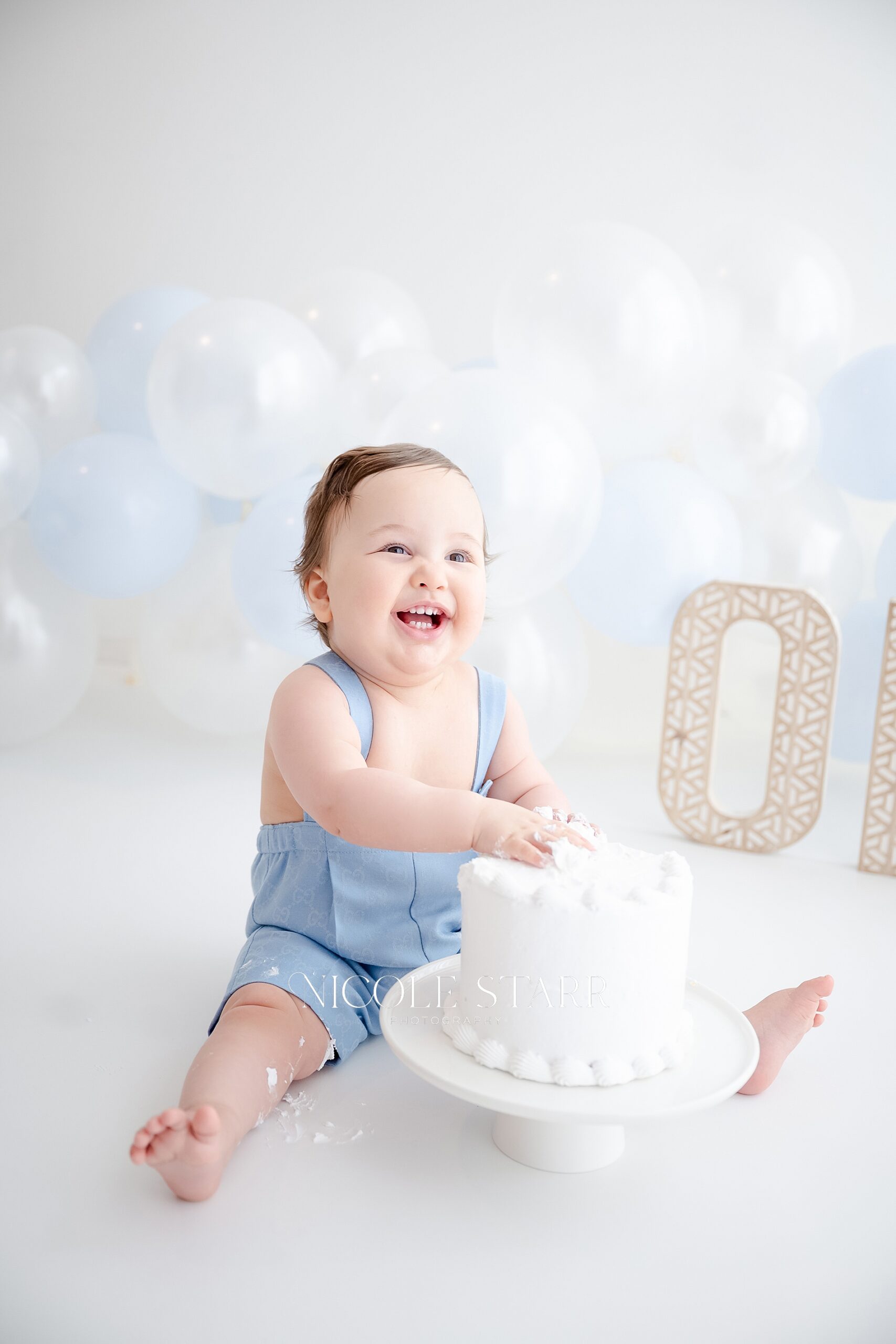 adorable one year old digs into white cake during classic blue and white cake smash for baby’s first birthday